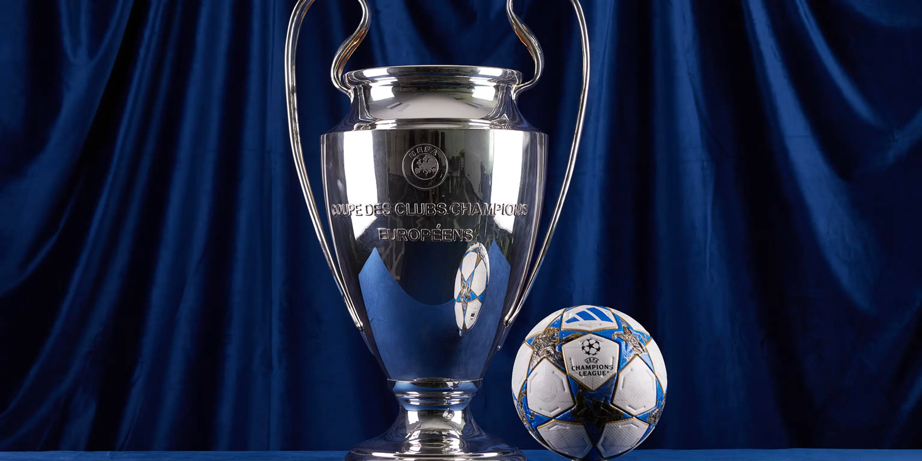The UEFA Champions League trophy beside an official Adidas match ball against a deep blue satin backdrop — representing the live European football coverage available across all devices on every IPTV subscription plan