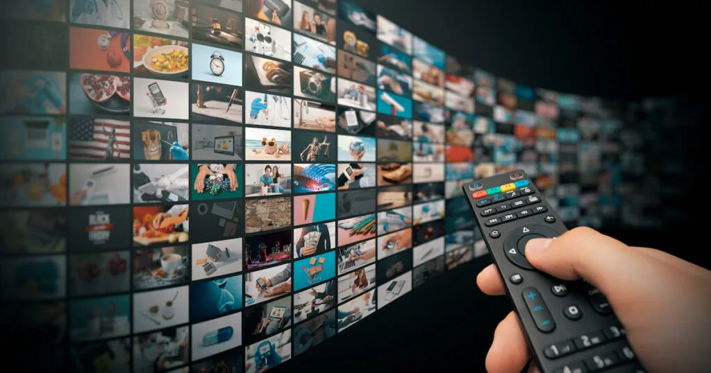 Hand holding a TV remote pointing at a massive wall of hundreds of streaming content channels — illustrating the breadth of programming available through a US IPTV service